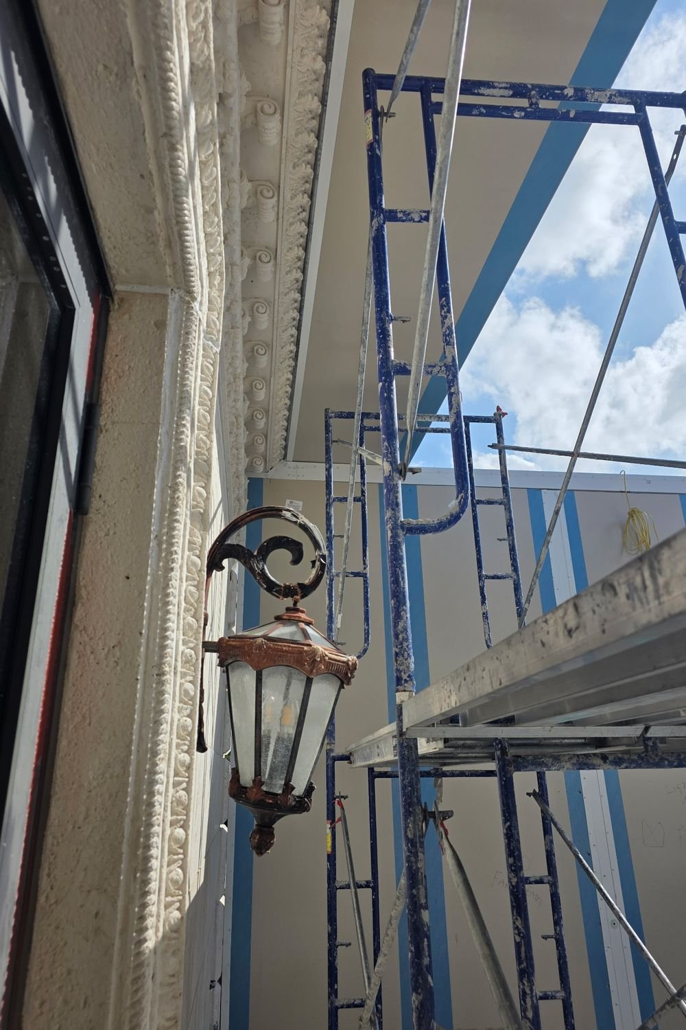 A bronze and copper carriage lantern mounted on an ornate cast stone facade, with scaffolding erected alongside it.