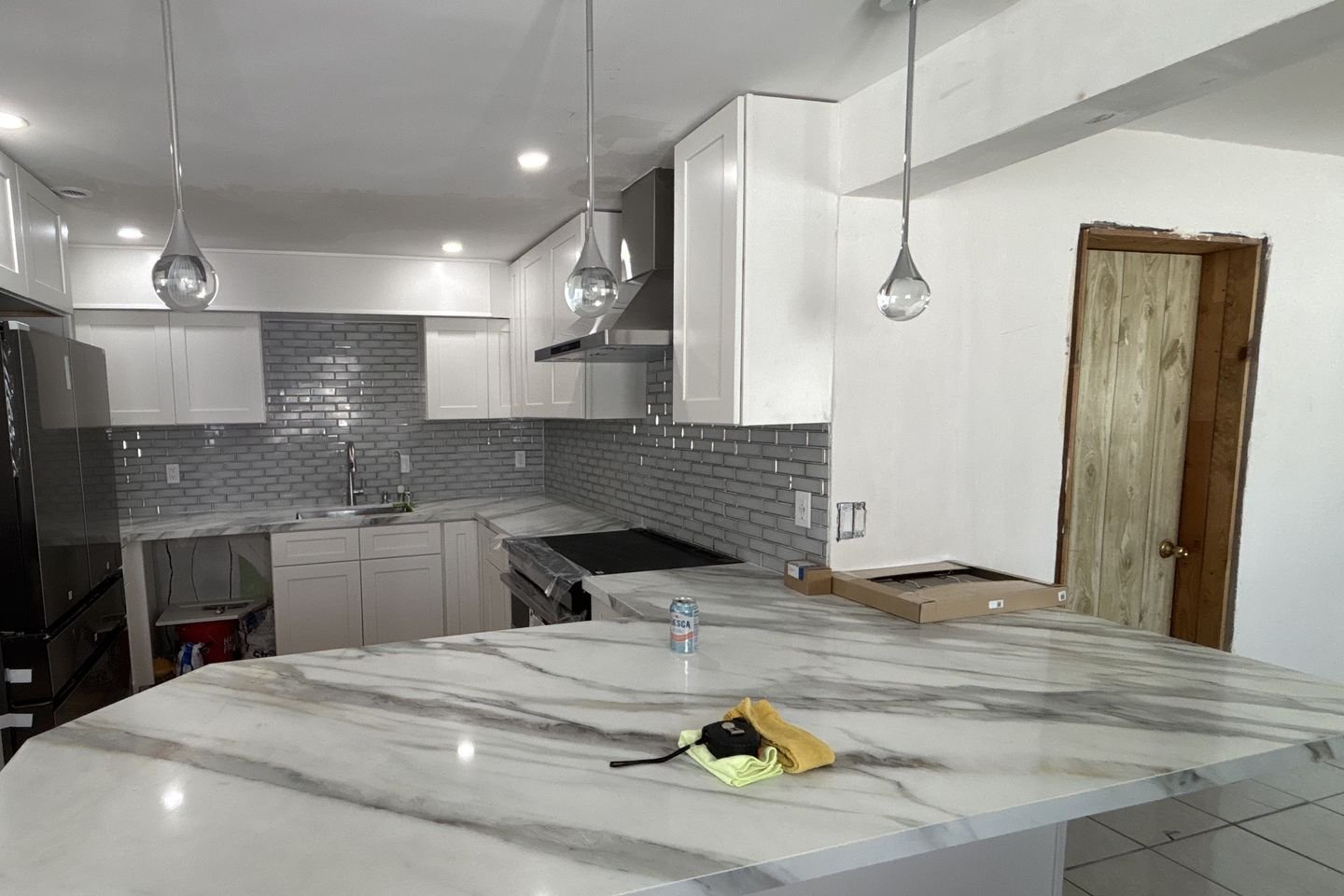The finished kitchen with white shaker cabinets, a stacked gray tile backsplash, chrome pendant lights and a veined stone peninsula.